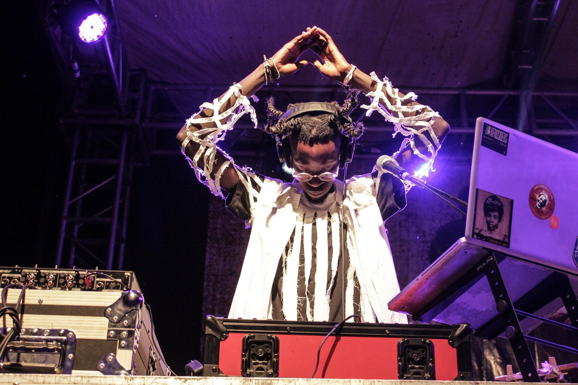 A performer in white and black making a heart with her hands above a DJ deck under purple stage light