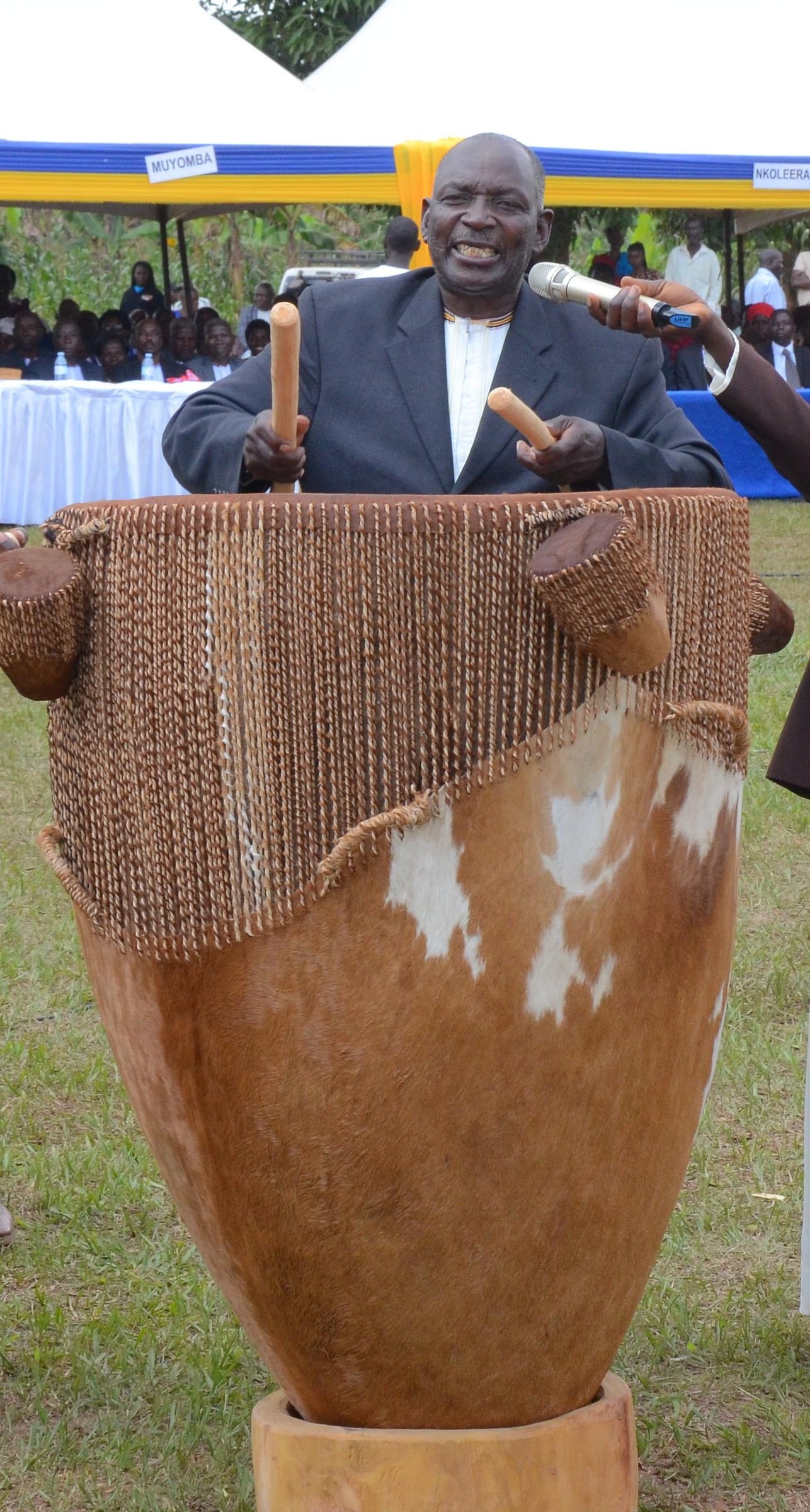 An elder playing a large cowhide drum with two sticks at an outdoor ceremony